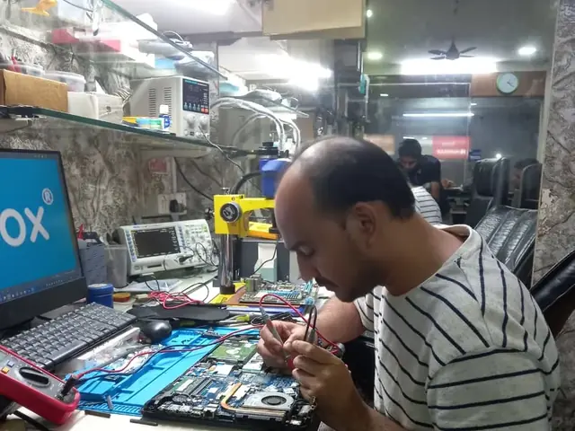 Technician testing a laptop mainboard with a multimeter beside a Xigox-branded monitor