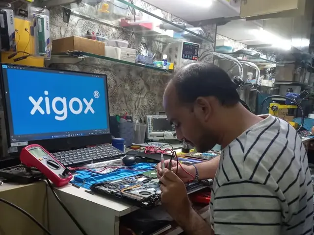 Technician testing a laptop mainboard with a multimeter beside a Xigox-branded monitor