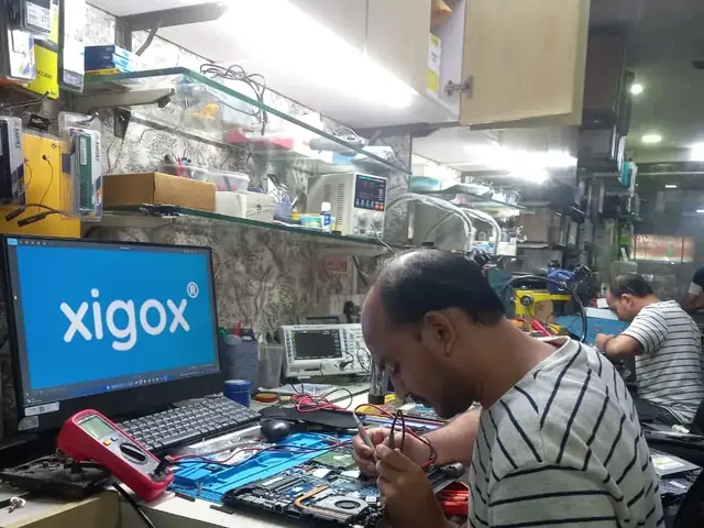 Technician testing a laptop mainboard with a multimeter beside a Xigox-branded monitor