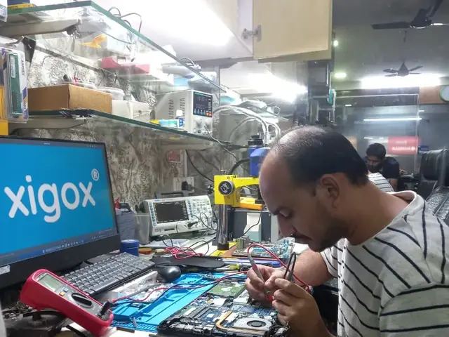 Technician testing a laptop mainboard with a multimeter beside a Xigox-branded monitor