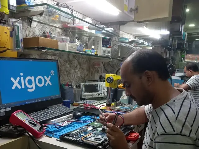 Technician testing a laptop mainboard with a multimeter beside a Xigox-branded monitor