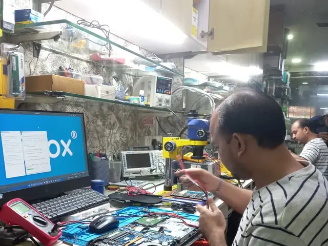 Technician testing a laptop mainboard with a multimeter beside a Xigox-branded monitor