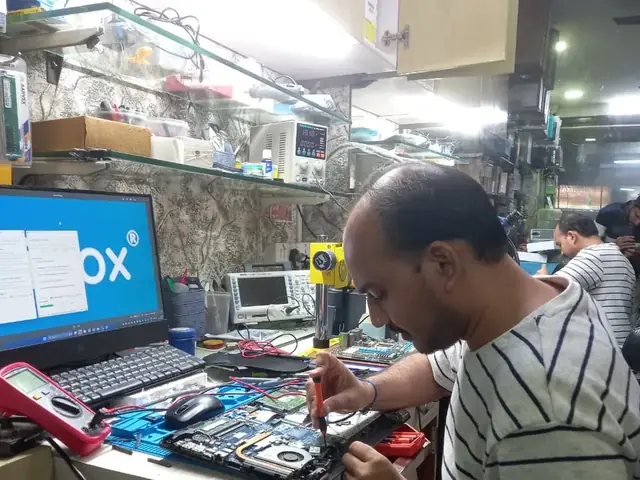 Technician testing a laptop mainboard with a multimeter beside a Xigox-branded monitor